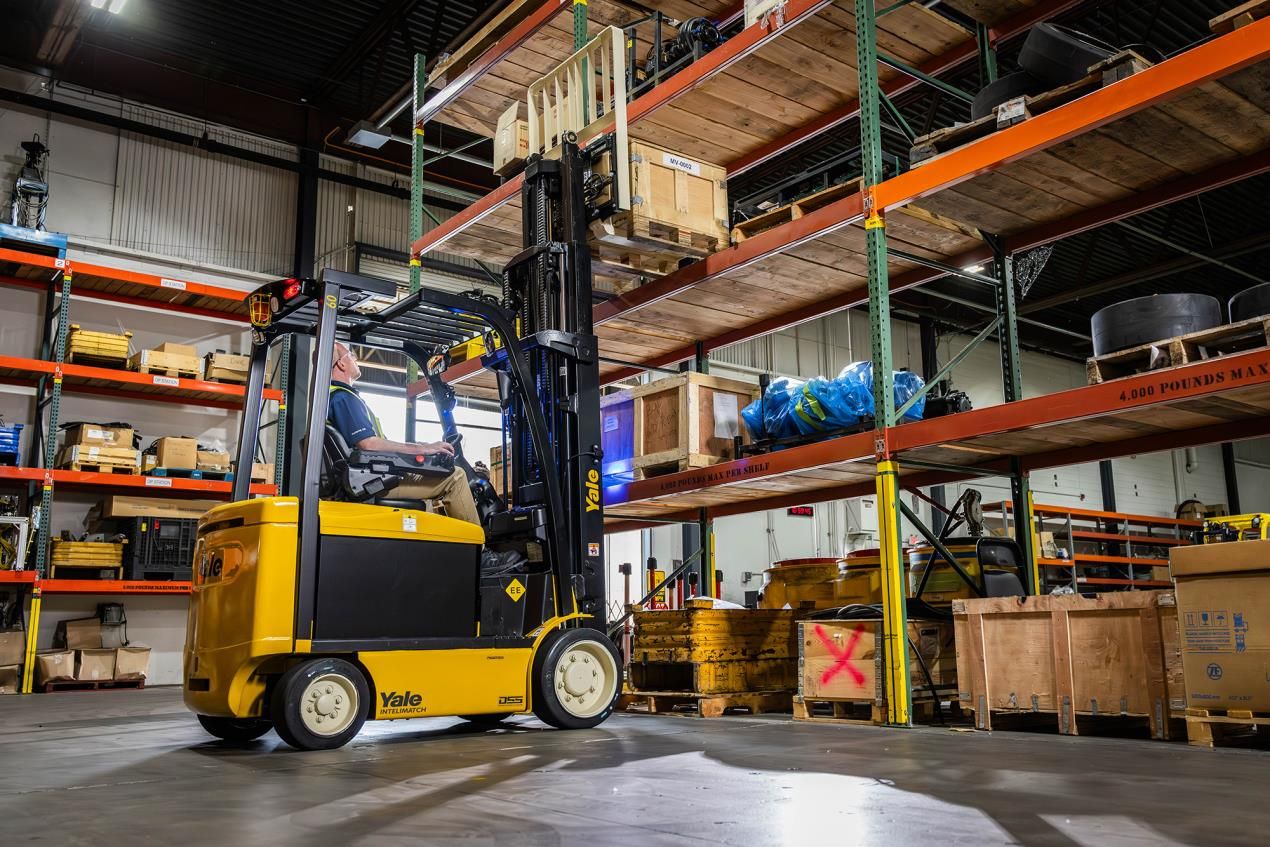 Yale electric lift truck with raised mast and load, seen side-on in a warehouse with pallet racking