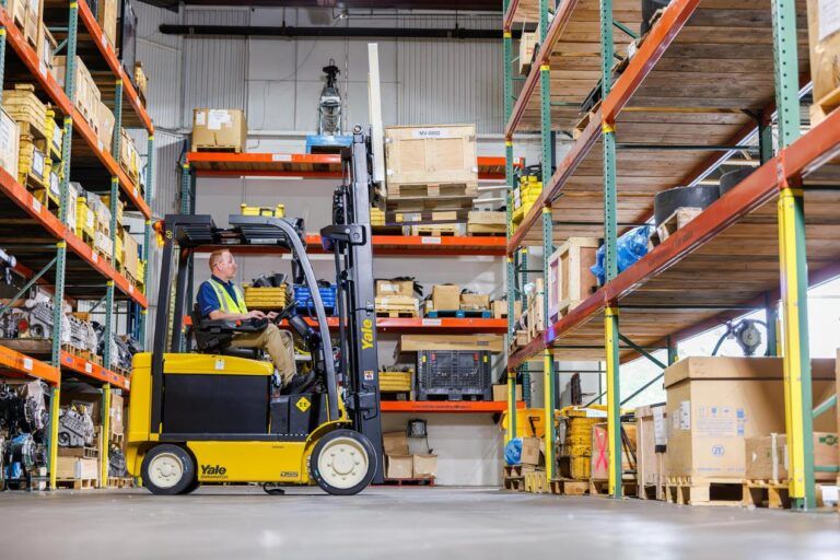 Yale updates flagship electric counterbalanced lift truck Yellow and black Yale electric lift truck with seated operator in a hi-vis vest, moving through a warehouse racking aisle