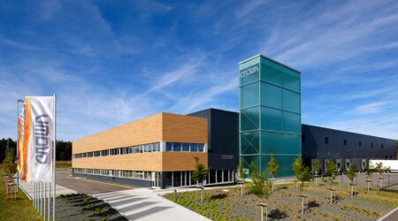 Crown marks 40 years of manufacturing at Roding Exterior view of Crown’s modern Roding manufacturing facility, a large building with timber-clad offices and a green glass tower, with Crown-branded flags in the foreground under a blue sky