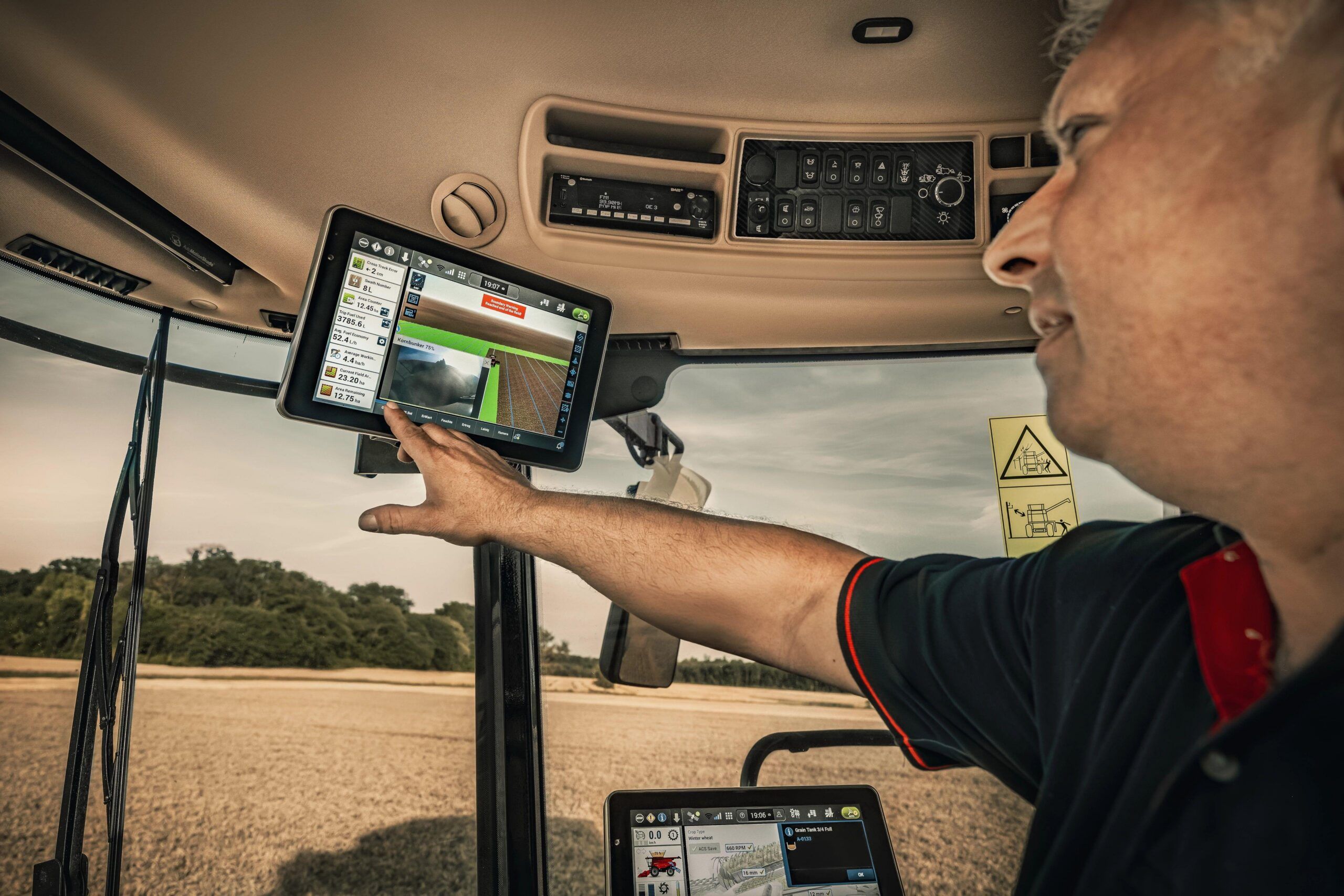An operator inside an Axial-Flow 160 series combine cab reaching up to a touchscreen display showing harvest data, with a second display mounted lower and a harvested field beyond the windscreen.