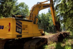 A Komatsu intelligent machine control excavator working an earth cut in a wooded area, digging a section of trail