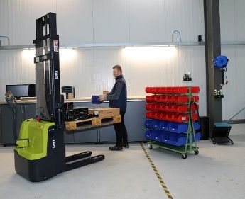 A green and black Clark electric high-lift pallet truck parked in a workshop beside a workbench and parts racking, with a worker handling a component in the background