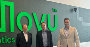 Three men in business dress stand in front of an illuminated green Movu Robotics wall sign