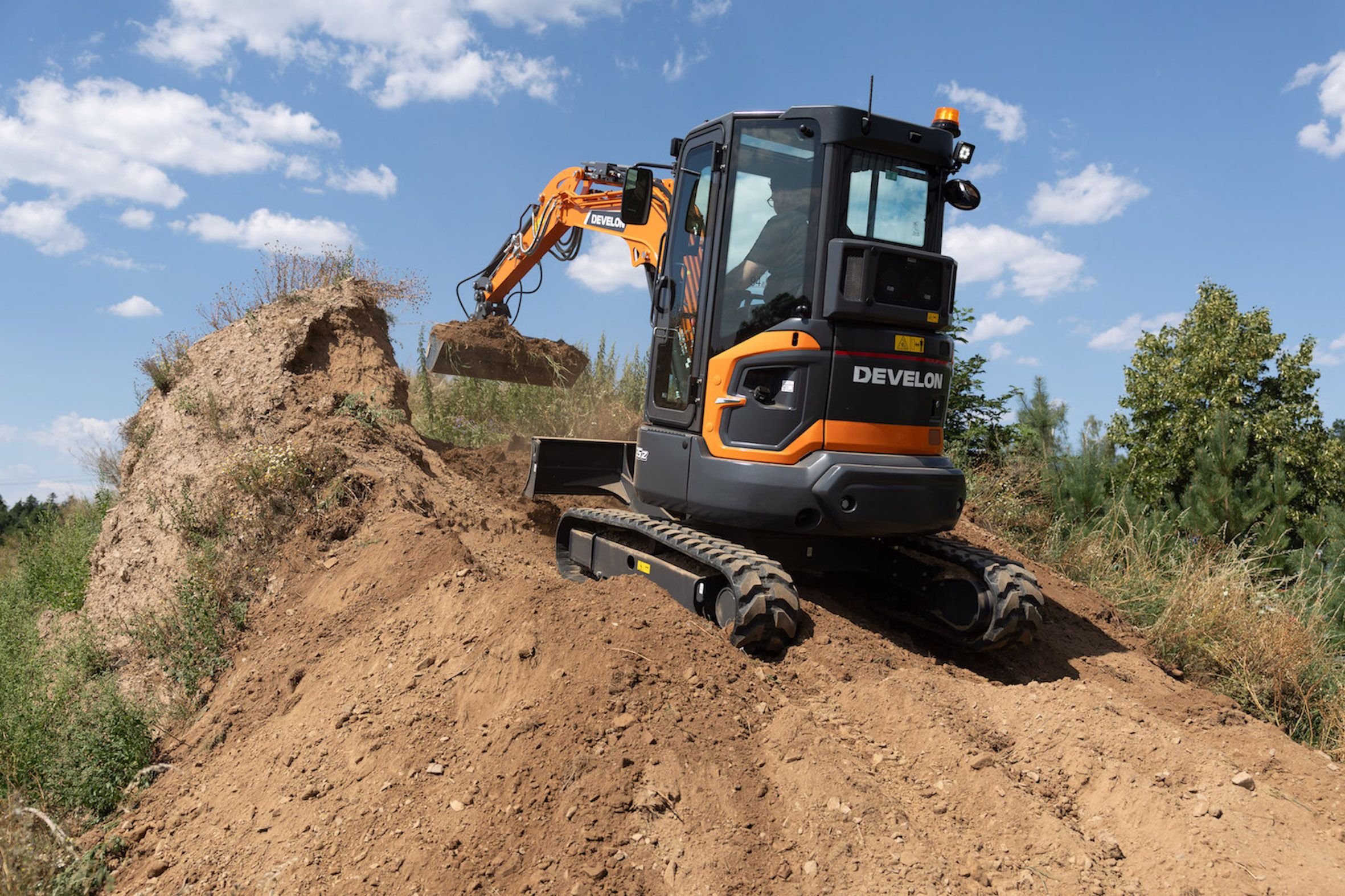 Rear three-quarter view of an orange Develon DX35Z-7 mini excavator working on top of a soil mound, showing the zero tail swing rear end and dozer blade.