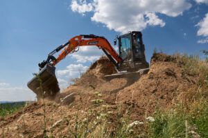 Orange Develon DX35Z-7 mini excavator digging into a soil mound on a hillside, bucket curled with soil spilling.