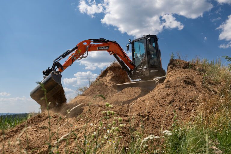 Develon upgrades DX35Z-7 mini excavator with new engine Orange Develon DX35Z-7 mini excavator digging into a soil mound on a hillside, bucket curled with soil spilling.