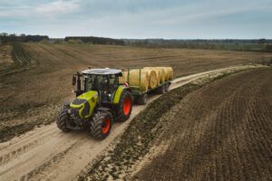 Aerial view of a green Claas Arion tractor towing a flatbed trailer loaded with round straw bales along a dirt track between ploughed fields.