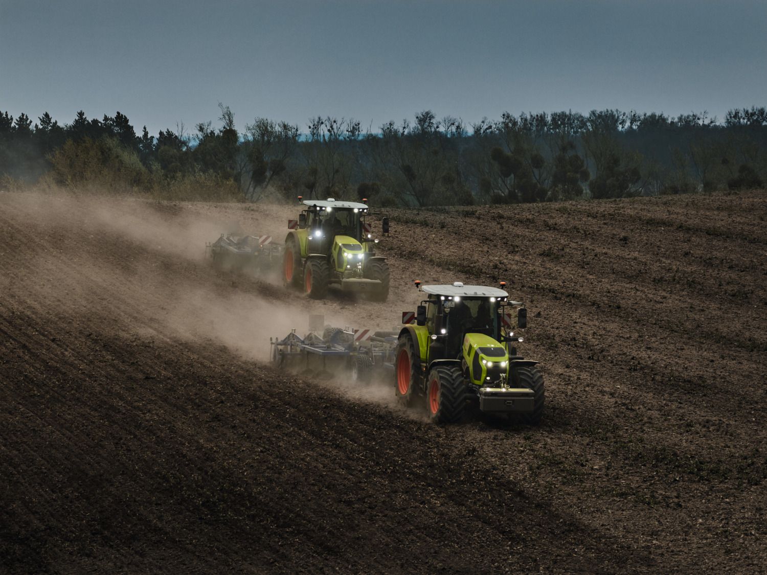 Two green Claas Arion tractors working a field at dusk with their headlights on, each drawing a cultivator and raising clouds of dust.