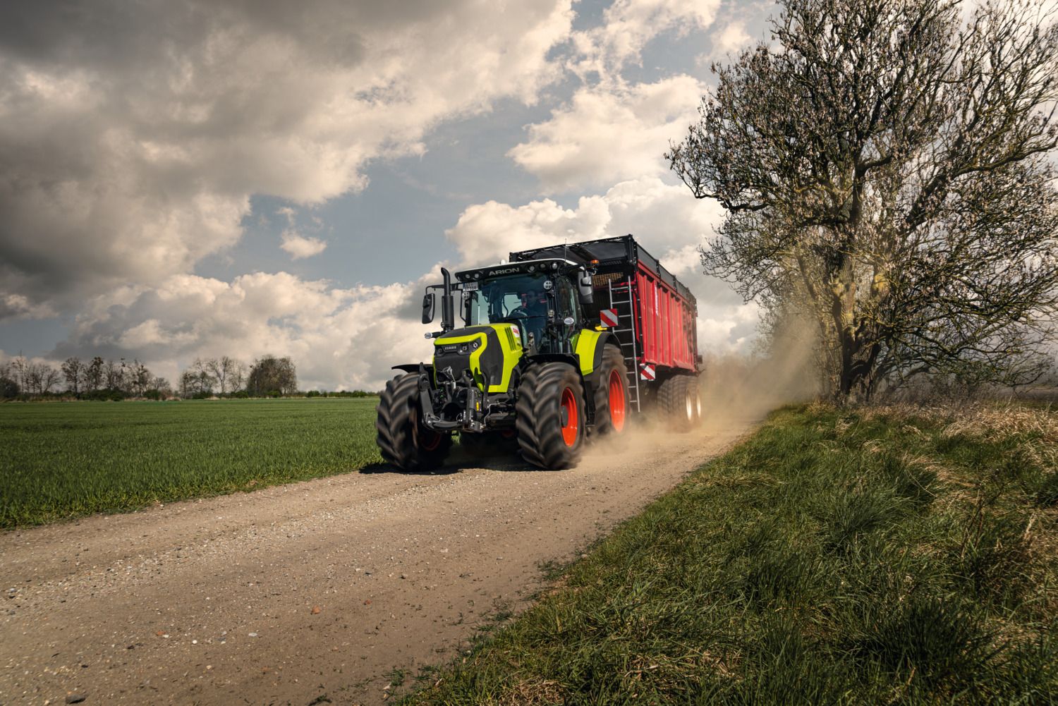 A green Claas Arion tractor towing a red forage trailer along a gravel farm track under a dramatic cloudy sky