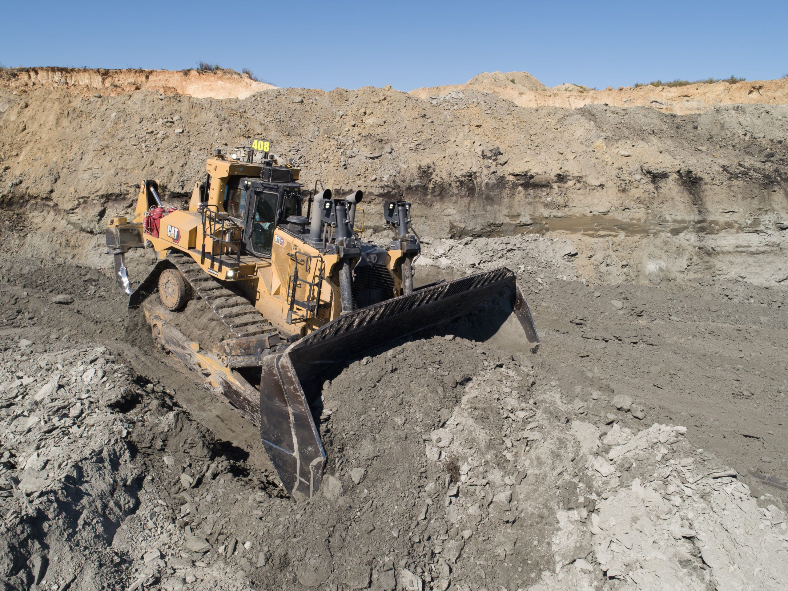 Elevated view of a Cat D11 XE dozer working a bench in an open pit, blade full of material, with excavated rock faces behind under a clear sky