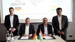 Four men in suits, two seated, sign an agreement with small German and Indian flags mounted on the desk foreground
