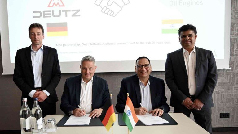 Four men in suits, two seated, sign an agreement with small German and Indian flags mounted on the desk foreground