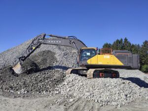 Volvo EC500 Electric crawler excavator with a yellow and grey body, digging into a large pile of grey crushed rock under a clear blue sky, with a cable reel mounted on its upper structure.