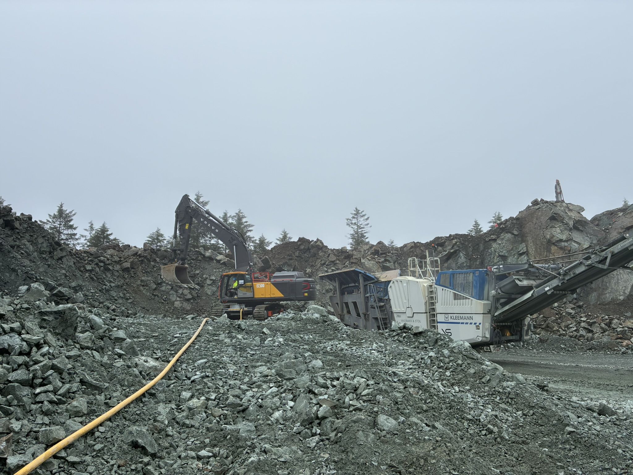 Volvo EC500 Electric excavator working in a misty quarry beside a white Kleemann mobile crushing plant, with a yellow power cable running across the crushed rock in the foreground.