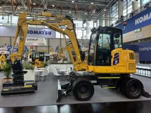 A yellow Komatsu PW75MR-5 compact wheeled excavator on an indoor exhibition stand, shown side-on with a front dozer blade, four road wheels and a boom-mounted working head