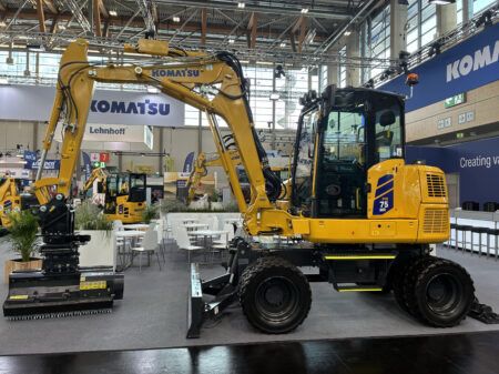 Komatsu Europe launches PW75MR-5 compact wheeled excavator A yellow Komatsu PW75MR-5 compact wheeled excavator on an indoor exhibition stand, shown side-on with a front dozer blade, four road wheels and a boom-mounted working head