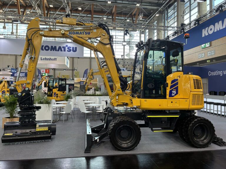 Komatsu Europe launches PW75MR-5 compact wheeled excavator A yellow Komatsu PW75MR-5 compact wheeled excavator on an indoor exhibition stand, shown side-on with a front dozer blade, four road wheels and a boom-mounted working head