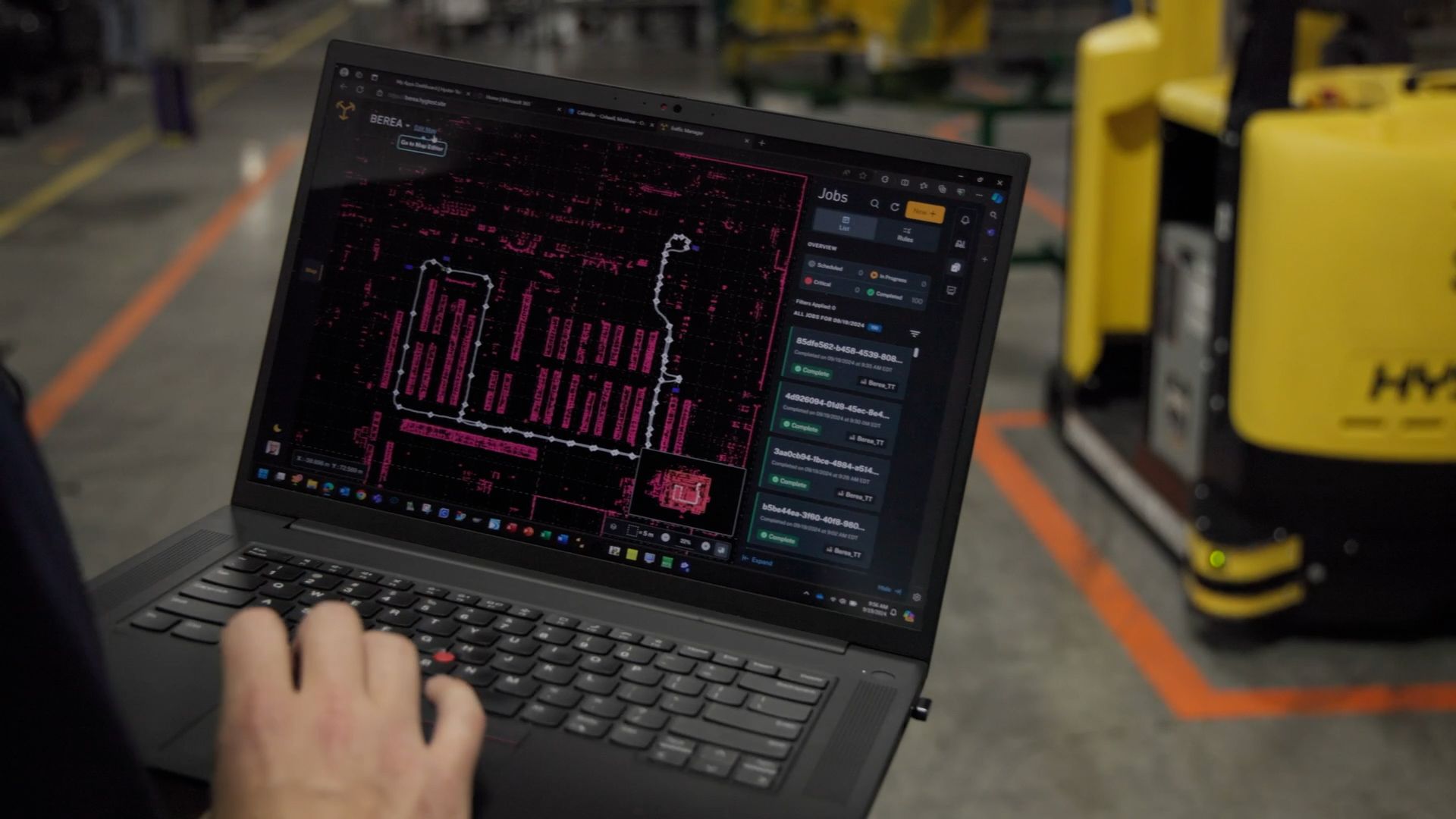 A laptop on a factory floor displays the Hyster Atlas portal, showing a pink facility map with a highlighted route and a Jobs panel listing completed tasks, with a yellow automated truck in the background.
