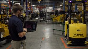 A Hyster employee holds a laptop showing the Atlas automation portal in a manufacturing aisle, with a yellow Hyster automated tow tractor numbered 399 standing on the floor beside marked lane lines.