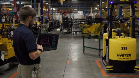 Hyster Atlas automation portal wins two product awards A Hyster employee holds a laptop showing the Atlas automation portal in a manufacturing aisle, with a yellow Hyster automated tow tractor numbered 399 standing on the floor beside marked lane lines.