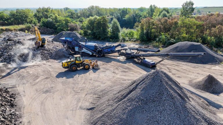 Blue Kleemann MOBIREX MR 130 EVO tracked mobile impact crusher operating on an outdoor recycling site, feeding a long tracked stacking conveyor with graded aggregate stockpiles alongside