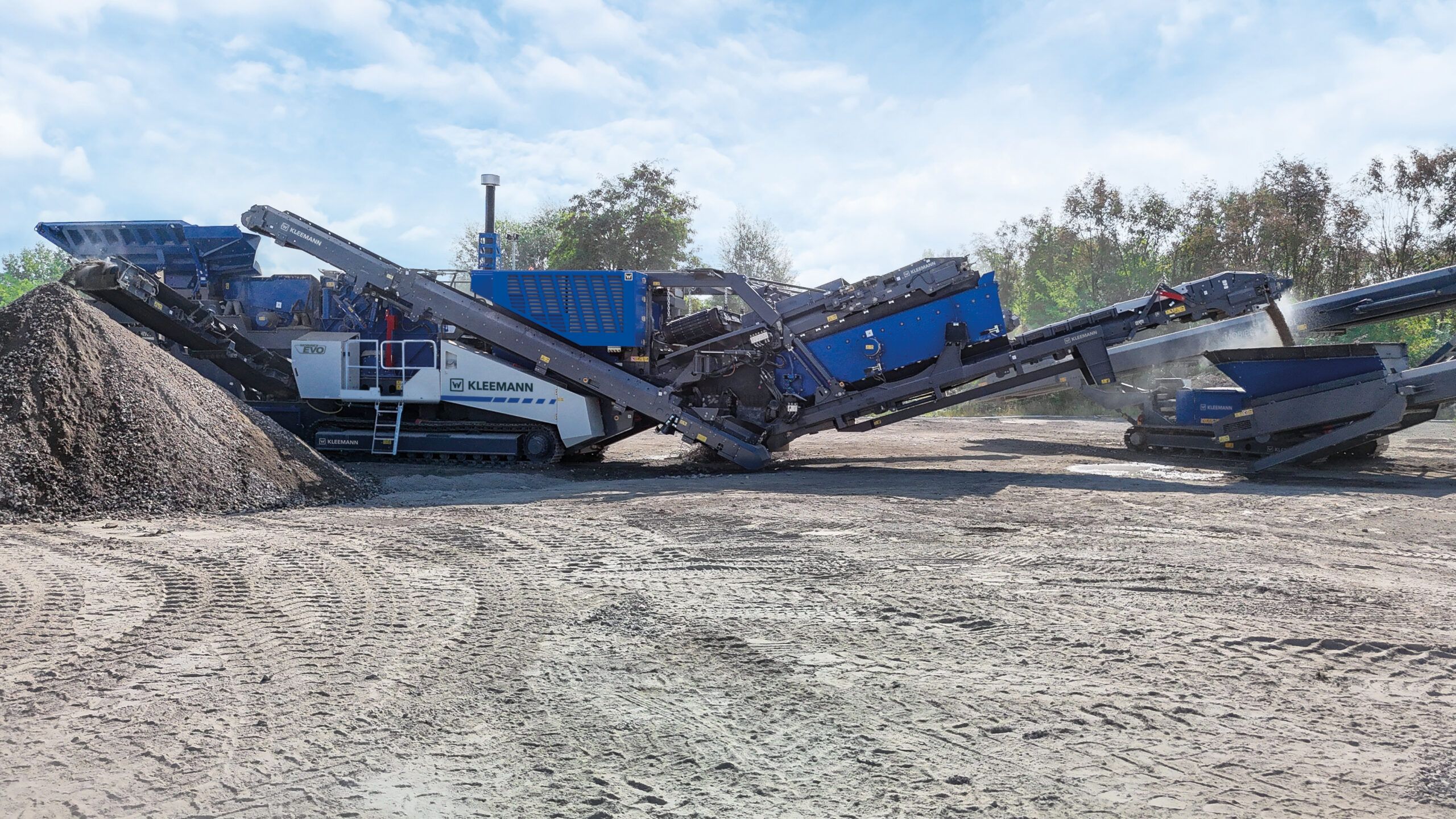 Side profile of the blue and white Kleemann MOBIREX MR 130 EVO tracked mobile impact crusher discharging crushed material onto a stockpile under a blue sky.