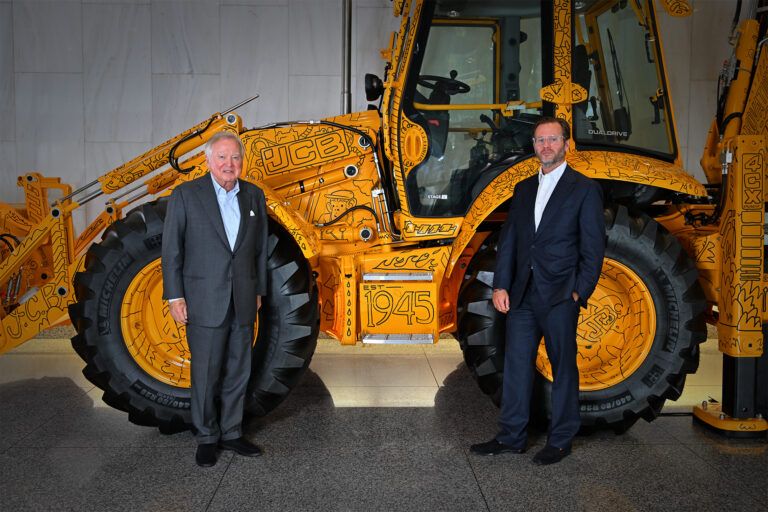 JCB names George Bamford joint chairman in first leadership change in 50 years Lord Bamford and George Bamford stand in front of a yellow JCB 4CX backhoe loader decorated with black line-drawing artwork and the wording EST 1945