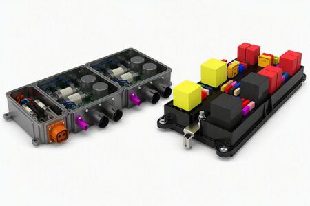 Two MTA power distribution units shown side by side against a white background: on the left, a grey Cube Box with its cover removed revealing internal circuitry and coloured connectors; on the right, a Modular High Voltage Box fitted with an array of yellow, red and multicoloured fuses and relays.