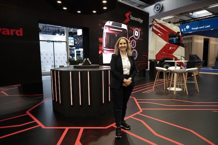 A woman in a dark blazer and white top stands smiling with her hands clasped in front of an exhibition stand, with a red-and-white liveried truck and screens displaying company branding in the background