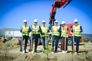 Six people in high-visibility vests and hard hats holding ceremonial shovels at a groundbreaking site, with a red Palfinger loader crane mounted on a truck behind them