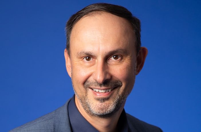 A head-and-shoulders portrait of a smiling man with short dark hair and grey stubble, wearing a grey blazer over a dark shirt, against a plain blue background