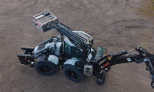 Aerial view of a white and black Huddig excavator loader on a gravel yard, with its front loader arm raised and a metal platform attachment lifted at the top, and its rear excavator arm folded