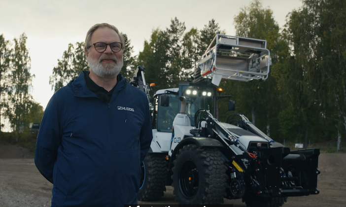 A man wearing a dark blue jacket with a Huddig logo stands in front of a Huddig excavator loader parked on a gravel site at dusk, with its headlights on and trees in the background.