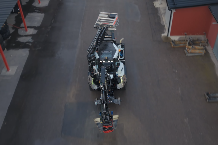 Aerial view looking straight down on a Huddig excavator loader driving along a paved yard between a building and a row of red bollards, with its arm and attachment extended at the front.