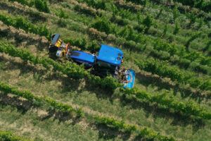 An overhead view of a blue New Holland specialty tractor working between rows of vines, with implements mounted front and rear