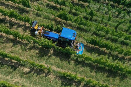 An overhead view of a blue New Holland specialty tractor working between rows of vines, with implements mounted front and rear