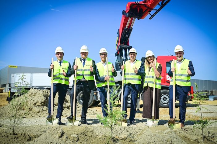 Six people in hard hats and high-visibility vests pose with golden shovels beside newly planted trees at the Palfinger groundbreaking ceremony, with a red crane truck behind them