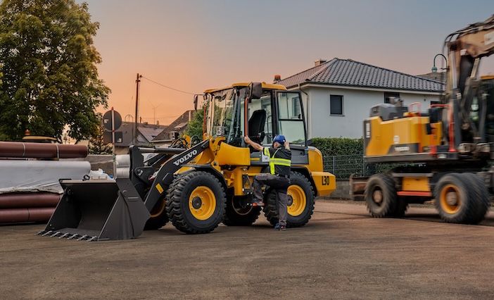 A yellow Volvo L30 wheel loader with a raised bucket is parked at a construction site at dusk, with an operator standing beside the cab, stacked pipes, a Volvo truck and an excavator on a trailer in the background, and houses beyond