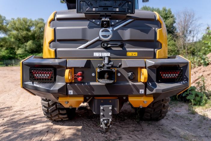 A close-up rear view of a yellow and grey wheel loader shows a central trailer hitch flanked by red and blue hydraulic quick-connect couplers, two grille-covered light clusters, and rugged bumper panels, with dirt ground and trees in the background