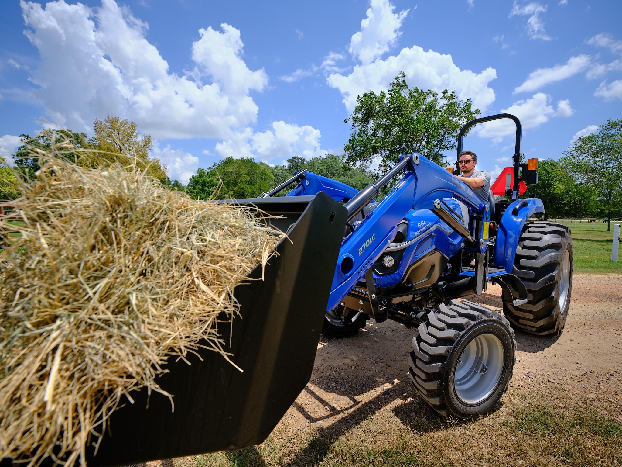 A blue New Holland Workmaster 55C compact tractor with an operator seated at the controls, using a 270LC front loader to carry a large round hay bale along a ranch track