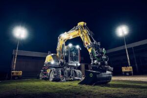 A yellow and black wheeled excavator with an attachment coupler stands on grass at night, lit by two tall floodlight towers with generator units positioned either side