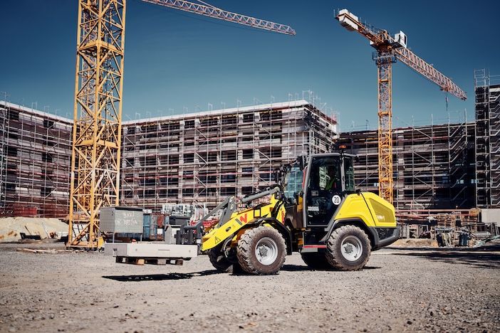 : A yellow wheel loader carries a pallet of concrete blocks on its front attachment across a construction site, with two tower cranes and a scaffolded building under construction behind it