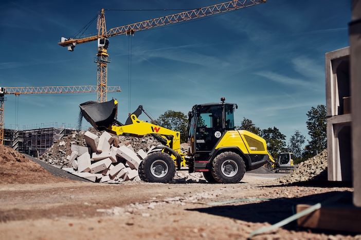 A yellow wheel loader lifts a bucket of demolition rubble at a construction site, with two tower cranes, scaffolded buildings and another machine visible in the background
