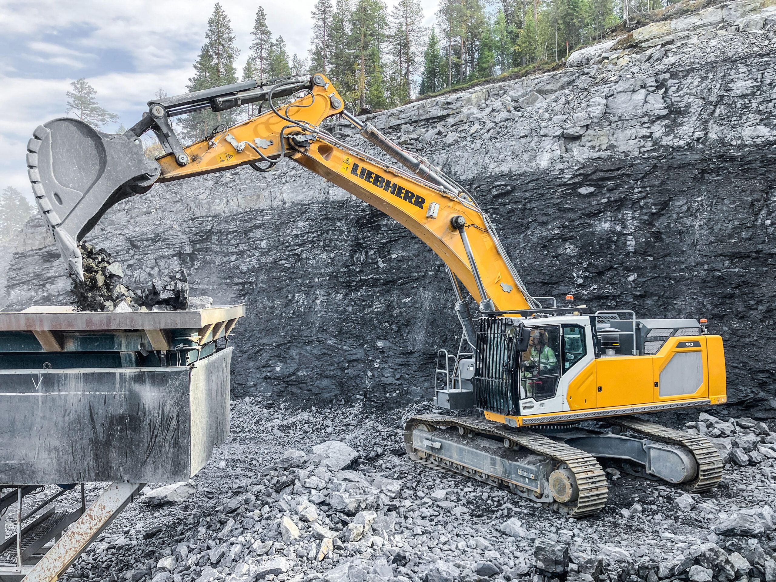 Yellow and white Liebherr R 952 G8 crawler excavator loading blasted rock into a truck body against a dark rock face in a cutting.