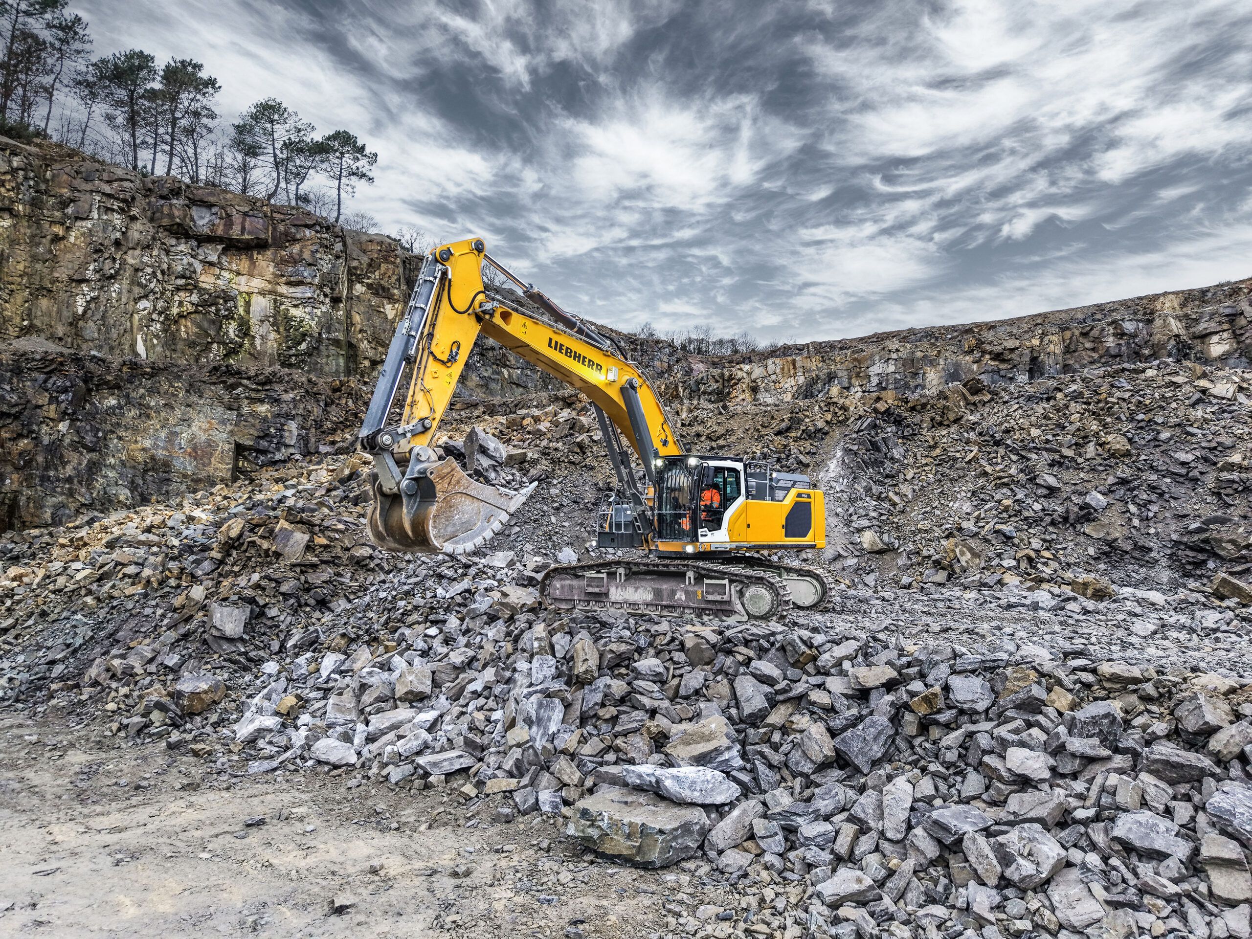 Yellow Liebherr R 958 G8 crawler excavator standing on broken stone in a quarry beneath a dramatic cloudy sky.