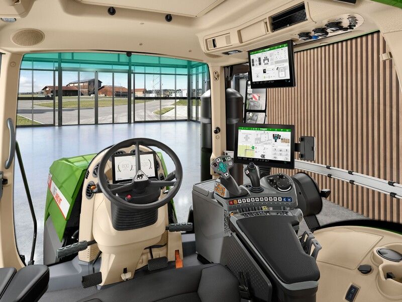 Interior of the Fendt 1100 Vario MT cab showing the steering wheel, digital dashboard, armrest terminal and roof-mounted display, with a showroom visible through the glass