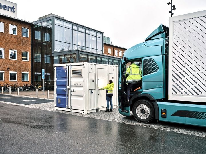 A teal-coloured electric truck is parked beside a white shipping container-style unit branded Volvo Penta, outside a brick and glass building; two people in high-visibility jackets stand at an open door on the unit, with bicycles parked in the background