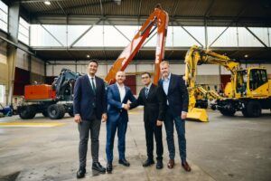 Four executives in business suits stand in an Atlas workshop, the two central figures shaking hands, with an orange Atlas material handler and yellow rail-mounted excavators behind them.