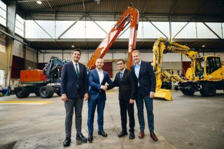 Buhler Versatile completes acquisition of Atlas Group operating business Four executives in business suits stand in an Atlas workshop, the two central figures shaking hands, with an orange Atlas material handler and yellow rail-mounted excavators behind them.