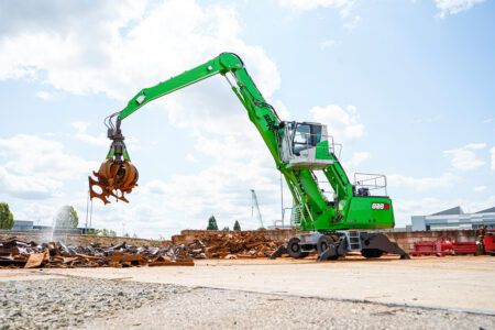 Sennebogen launches 828 G material handler A green Sennebogen 828 G material handler on a wheeled mobile undercarriage with its outriggers lowered, lifting scrap metal with an orange peel grab at a recycling yard under a bright sky, the 828 G badge visible on the green counterweight.